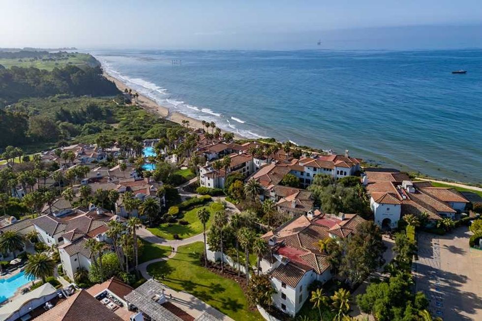 Coastal resort with palm trees, villas, and pools by a sandy beach and blue ocean.
