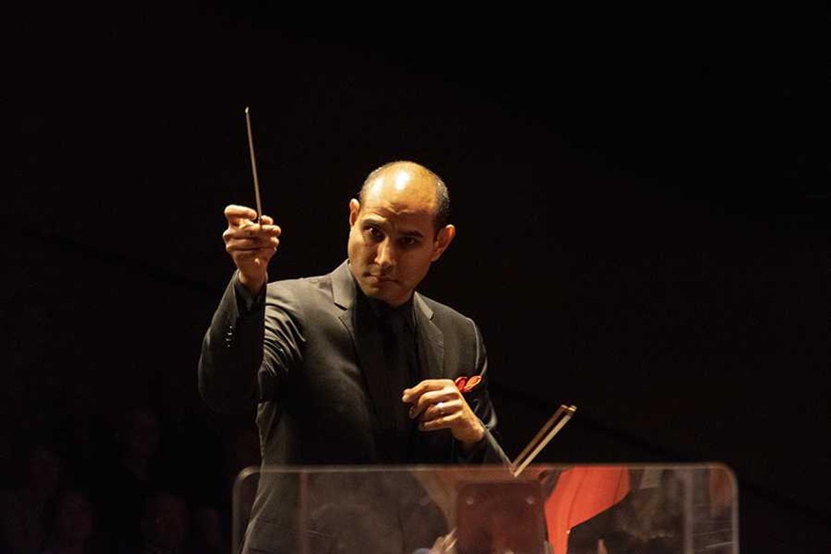 Conductor in a dark suit leading an orchestra in dim lighting.