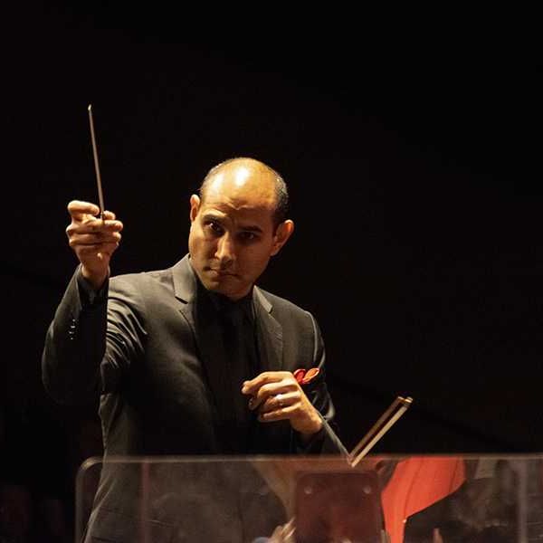 Conductor in a dark suit leading an orchestra in dim lighting.
