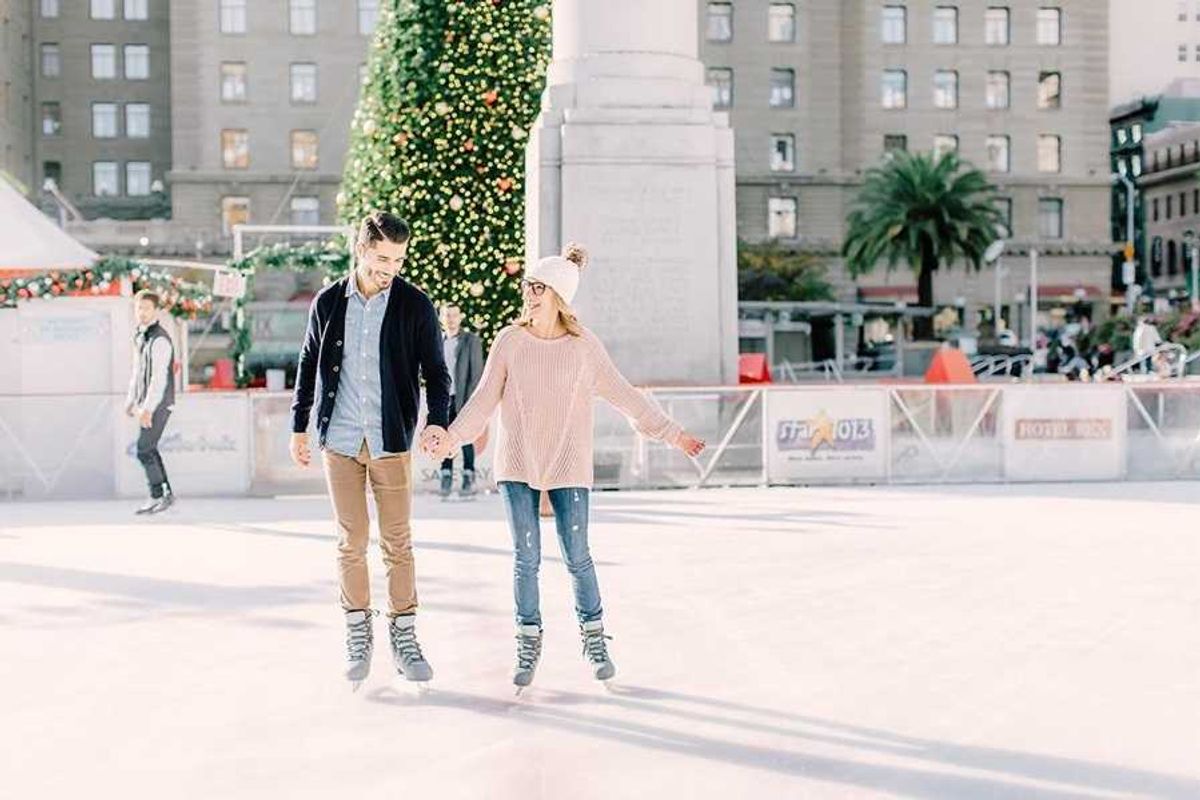 Couple ice skating near a decorated Christmas tree in a city square.