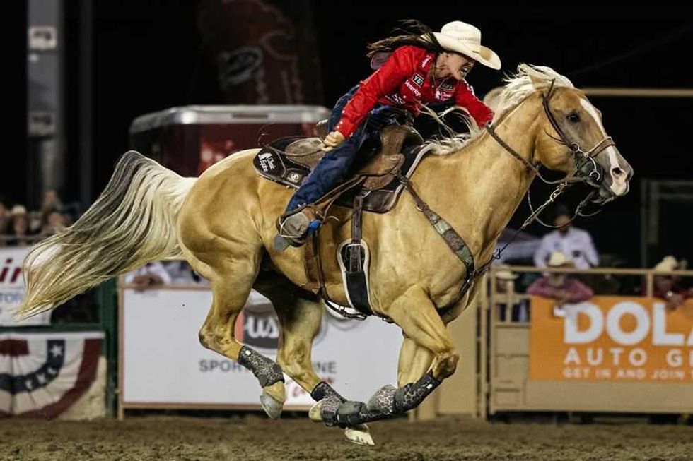 Cowgirl in red shirt racing palomino horse at nighttime rodeo event.