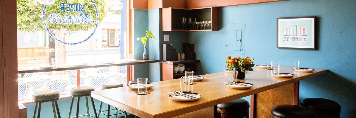 Cozy, modern restaurant interior with wooden table and stools by a large window.