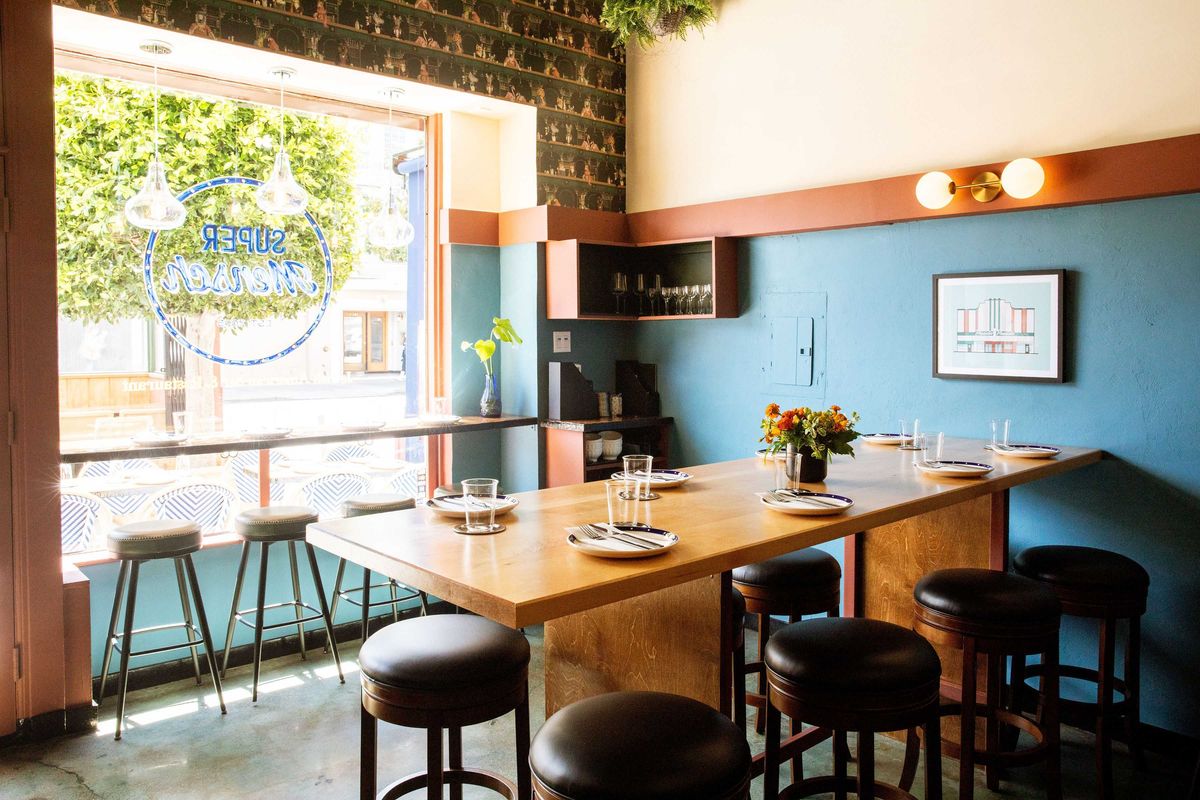Cozy, modern restaurant interior with wooden table and stools by a large window.