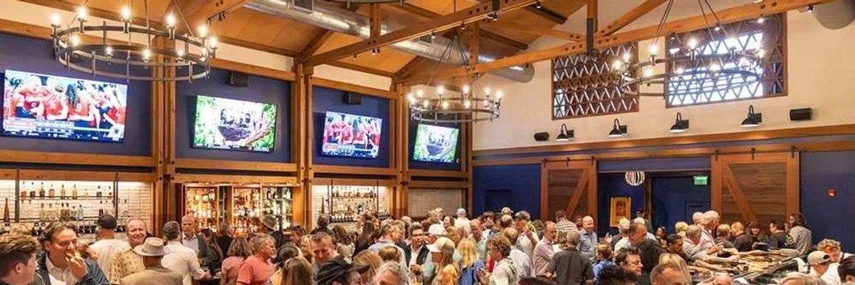 Crowded bar with people socializing under chandeliers and TV screens showing sports.