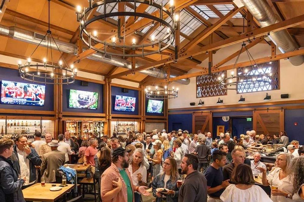 Crowded bar with people socializing under chandeliers and TV screens showing sports.