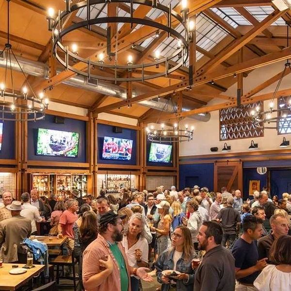Crowded bar with people socializing under chandeliers and TV screens showing sports.
