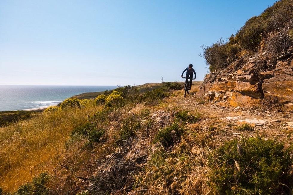 Cyclist riding a rocky coastal trail under a clear blue sky.