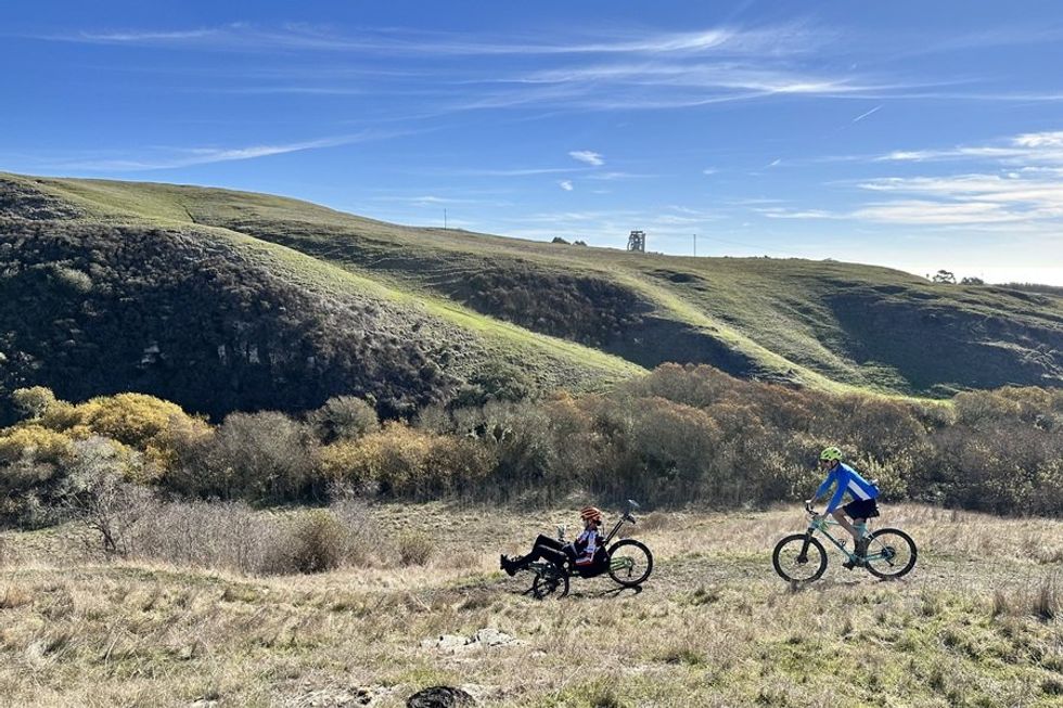 Cyclists on grassy trail with rolling hills and blue sky in background.