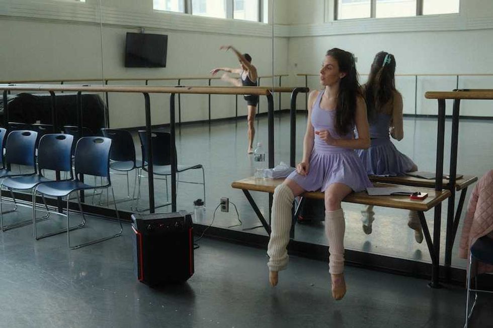 Dancer in a lilac outfit sits on a barre; another dancer moves in the mirrored background.