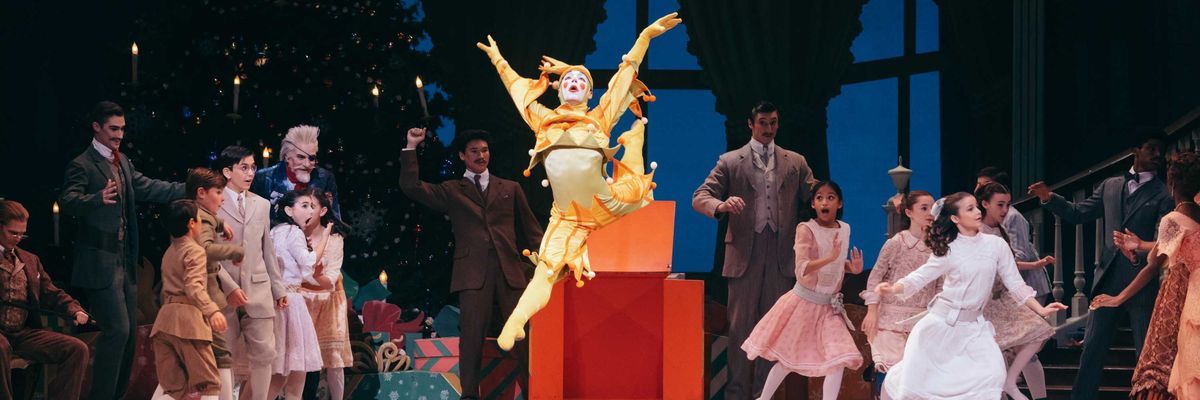 Dancer in vibrant costume leaps during a festive performance with a Christmas tree backdrop.