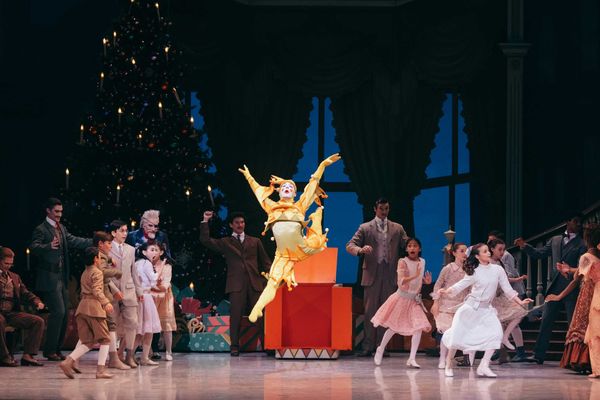 Dancer in vibrant costume leaps during a festive performance with a Christmas tree backdrop.