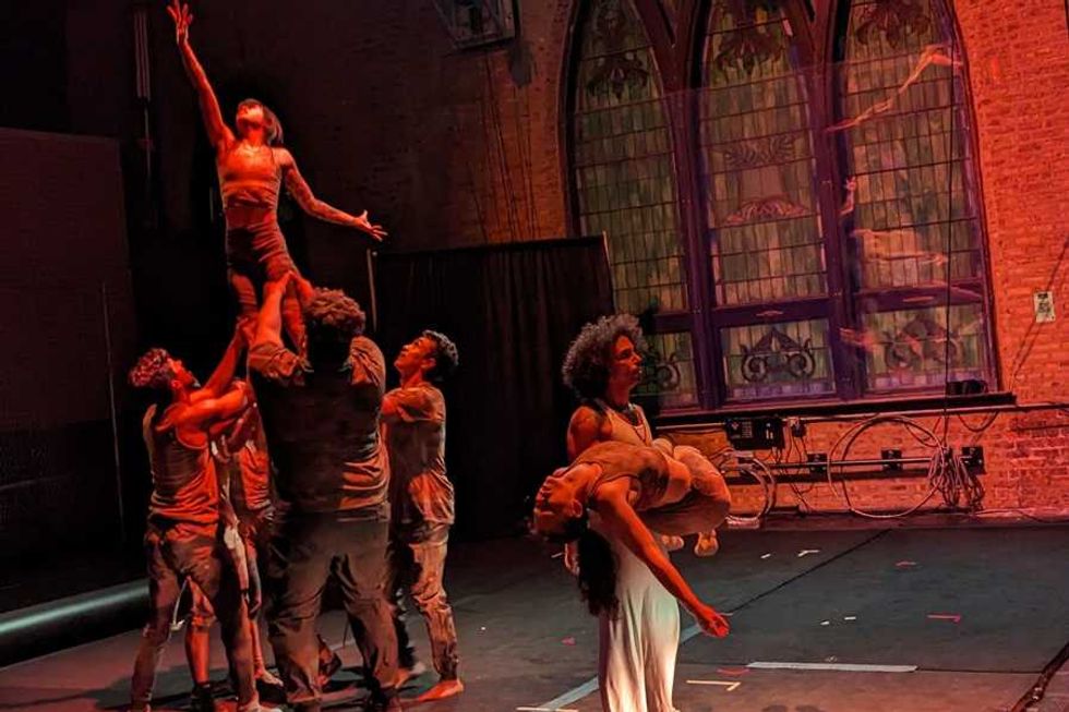 Dancers form a lifting pyramid while another holds a resting performer on a dimly lit stage.