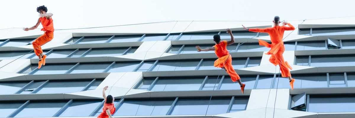Dancers in orange perform on a building facade, appearing to defy gravity.