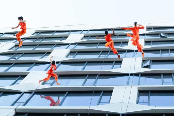 Dancers in orange perform on a building facade, appearing to defy gravity.