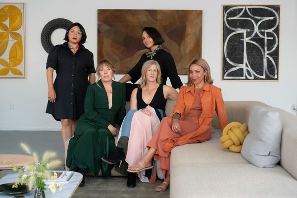 Five women pose in a stylish, modern living room with abstract art.