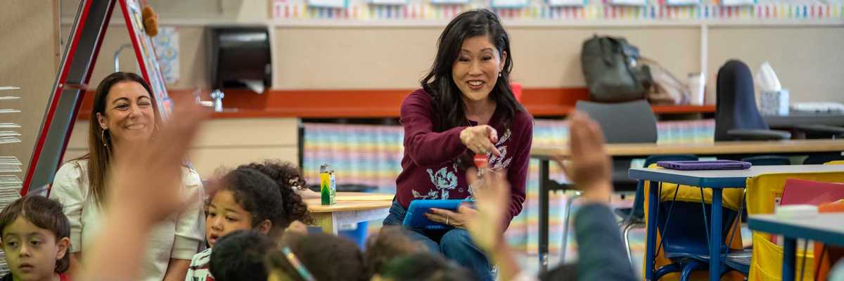 former ice skating champion Kristi Yamaguchi at the front of a kindergarten class pointing at a kid with a raised arm
