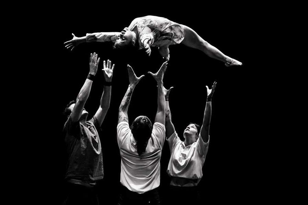 Four acrobats lifting a performer in mid-air against a black background.