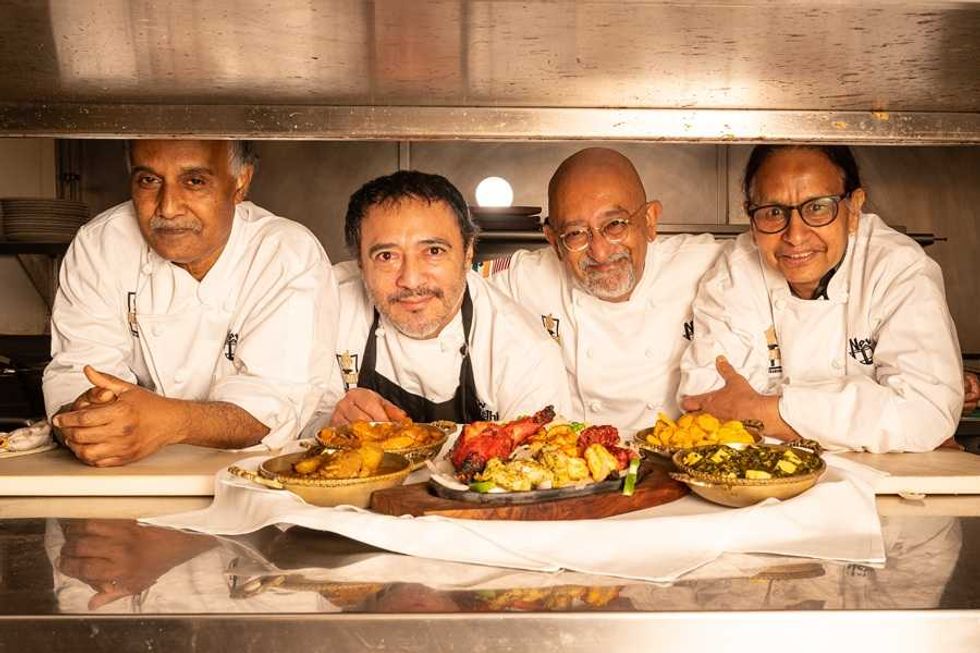Four chefs in white uniforms behind dishes of colorful food in a kitchen.