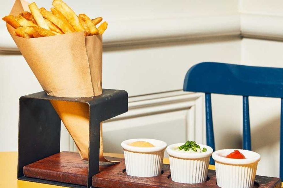 Fries in a metal holder with three dipping sauces, beside a blue chair.