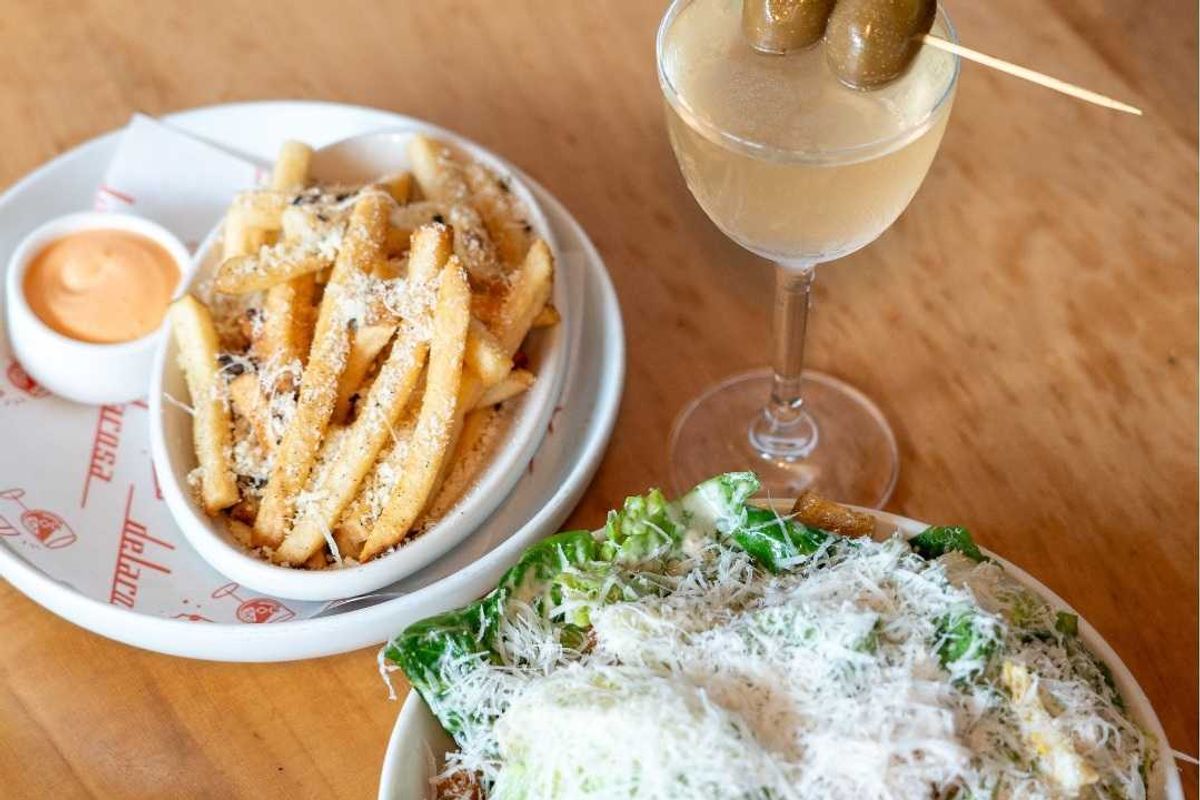 Fries with sauce, Caesar salad, and a cocktail with olives on a wooden table.