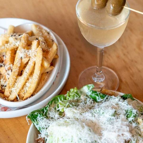 Fries with sauce, Caesar salad, and a cocktail with olives on a wooden table.