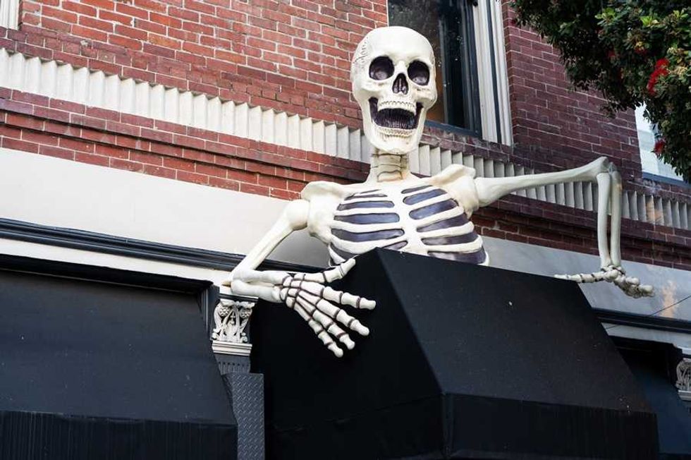 Giant skeleton on a roof, leaning on a brick building, with open mouth and outstretched arms.