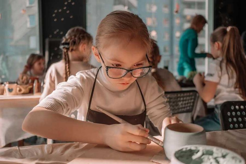 Girl in glasses painting pottery with a brush, focused in a classroom setting.