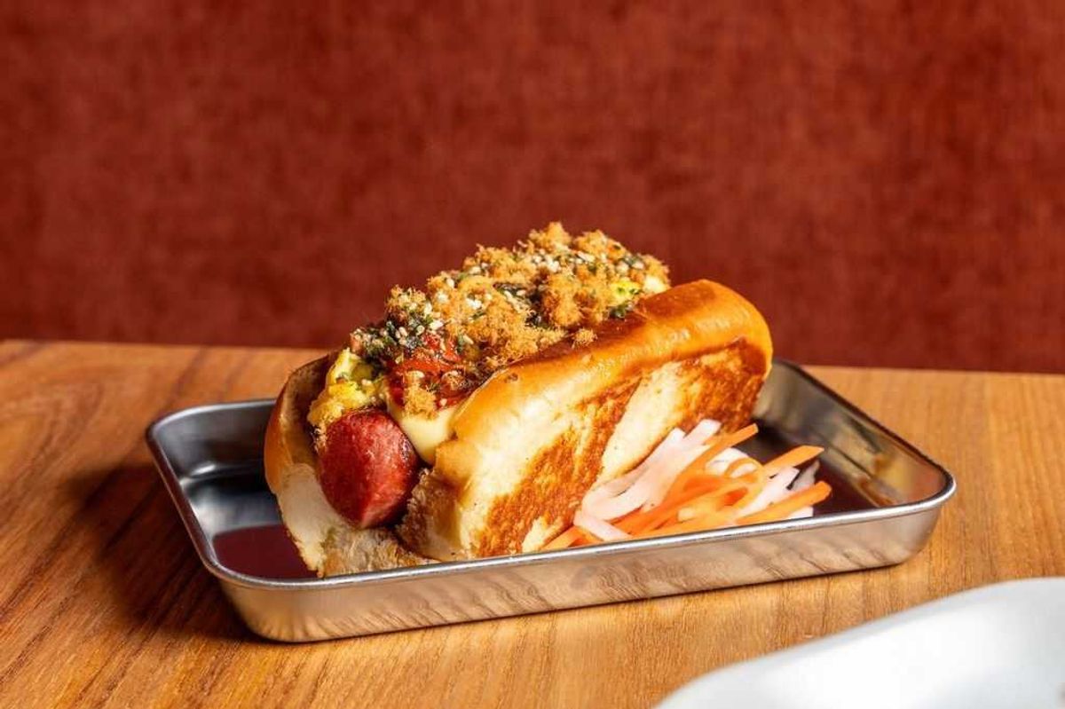 Gourmet hot dog with crumbled toppings in a tray, served on a wooden table.