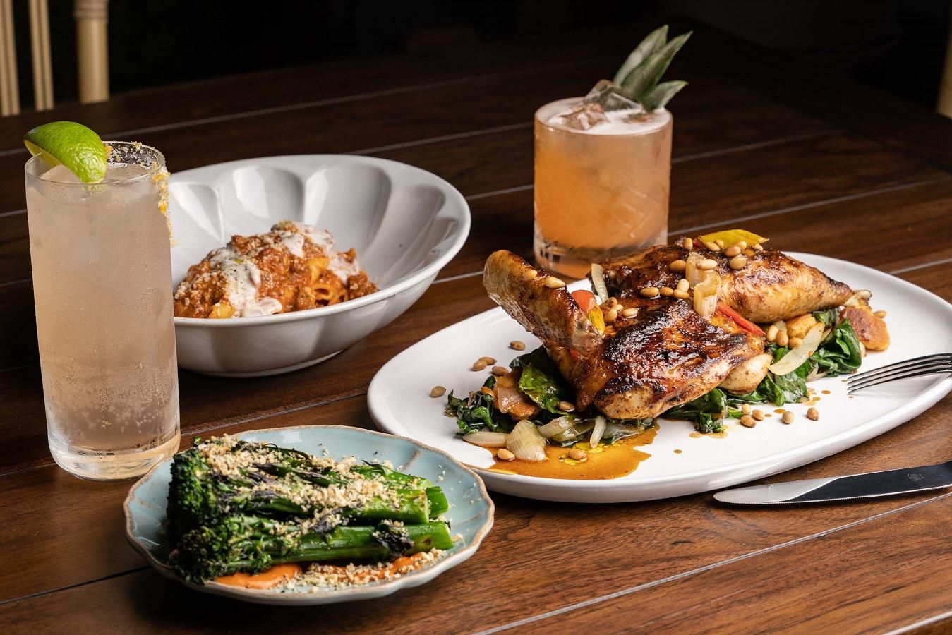 Gourmet meal with roast chicken, pasta, greens, and two cocktails on a wooden table.