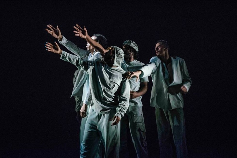 Group of dancers in white outfits reaching forward on a dark stage.