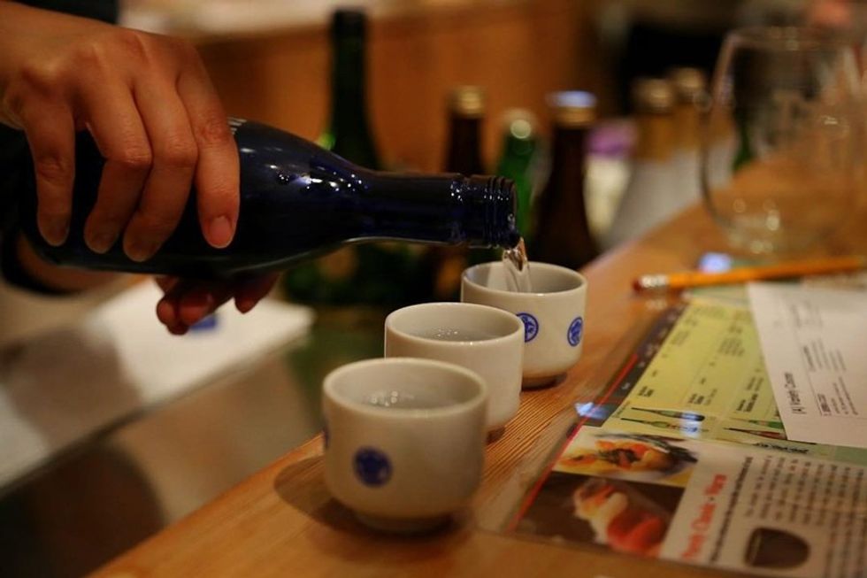 Hand pouring sake into small cups on a wooden counter with menus.