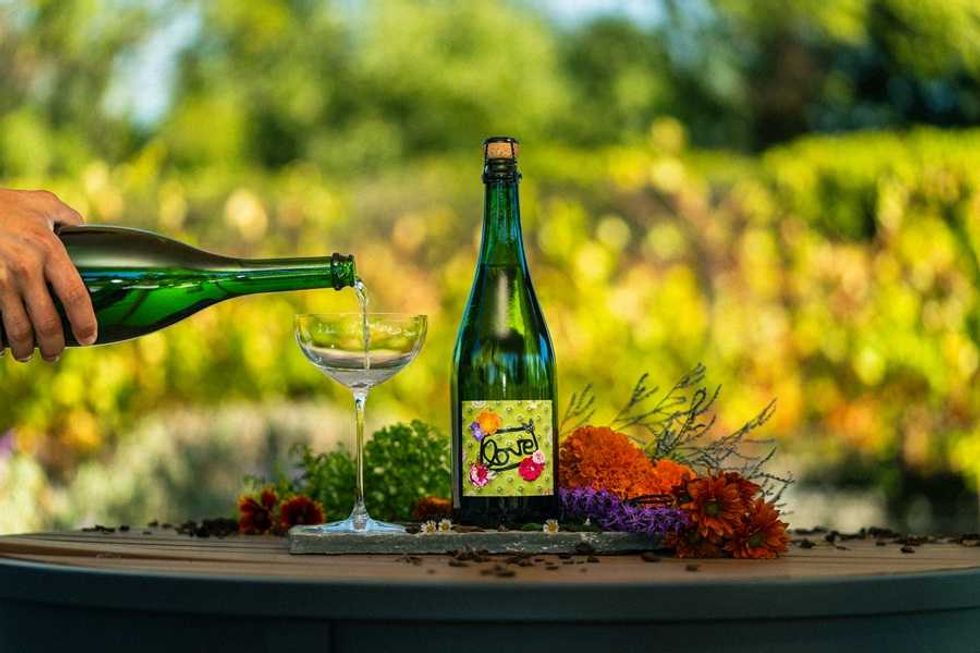 Hand pouring wine into a glass outdoors, bottle labeled "Love" on a flower-decorated table.