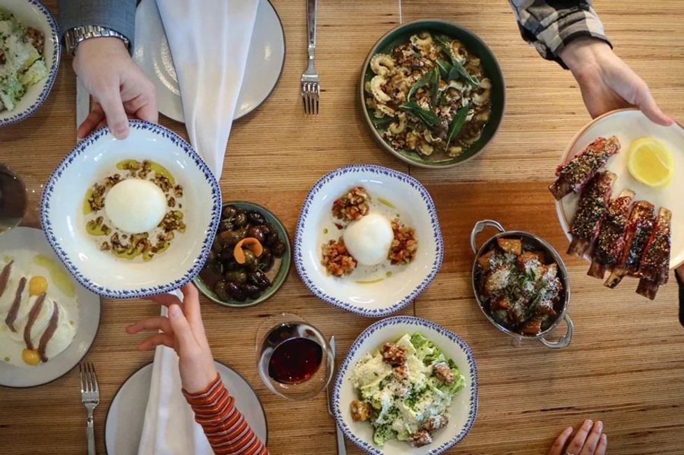 Hands passing and serving a variety of dishes on a wooden table.
