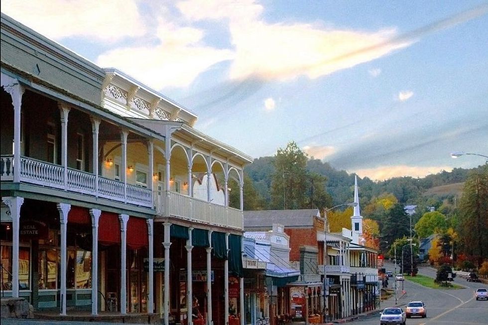 Historic street with colorful awnings and white church against a wooded hillside backdrop.