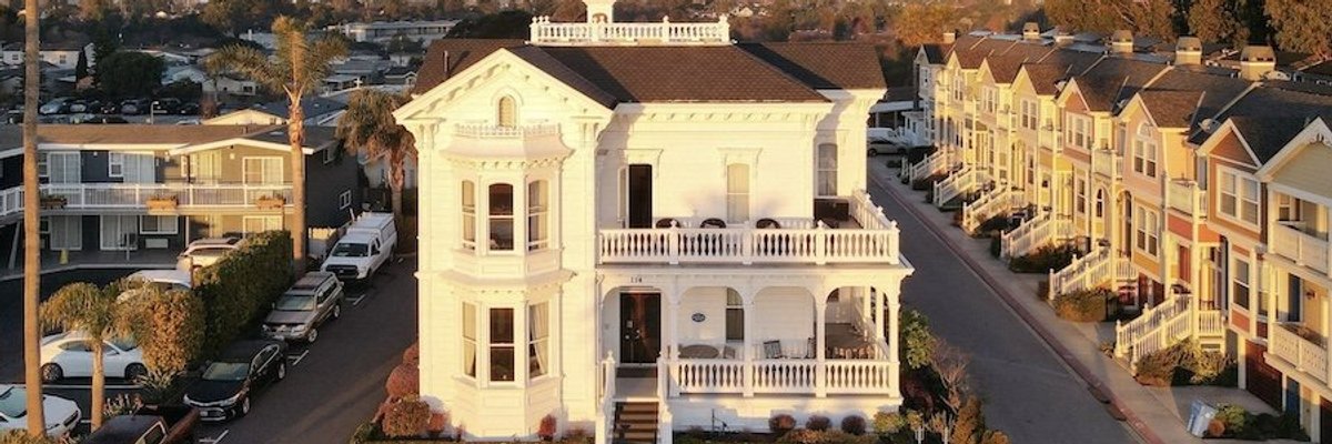 Historic white mansion with wraparound porch in a suburban neighborhood at sunset.