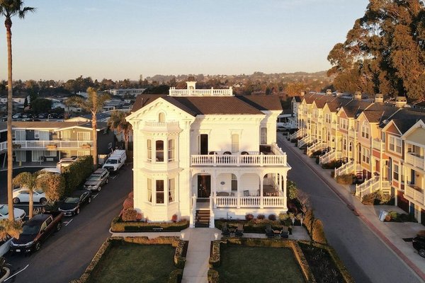 Historic white mansion with wraparound porch in a suburban neighborhood at sunset.