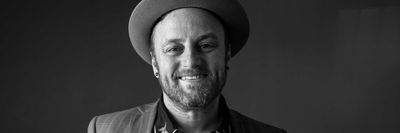 Smiling man in a hat and patterned shirt, wearing a plaid jacket, in black and white.