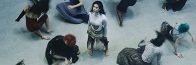 Aerial view of dancers in motion, surrounding one standing still, with blurred dynamic movements.