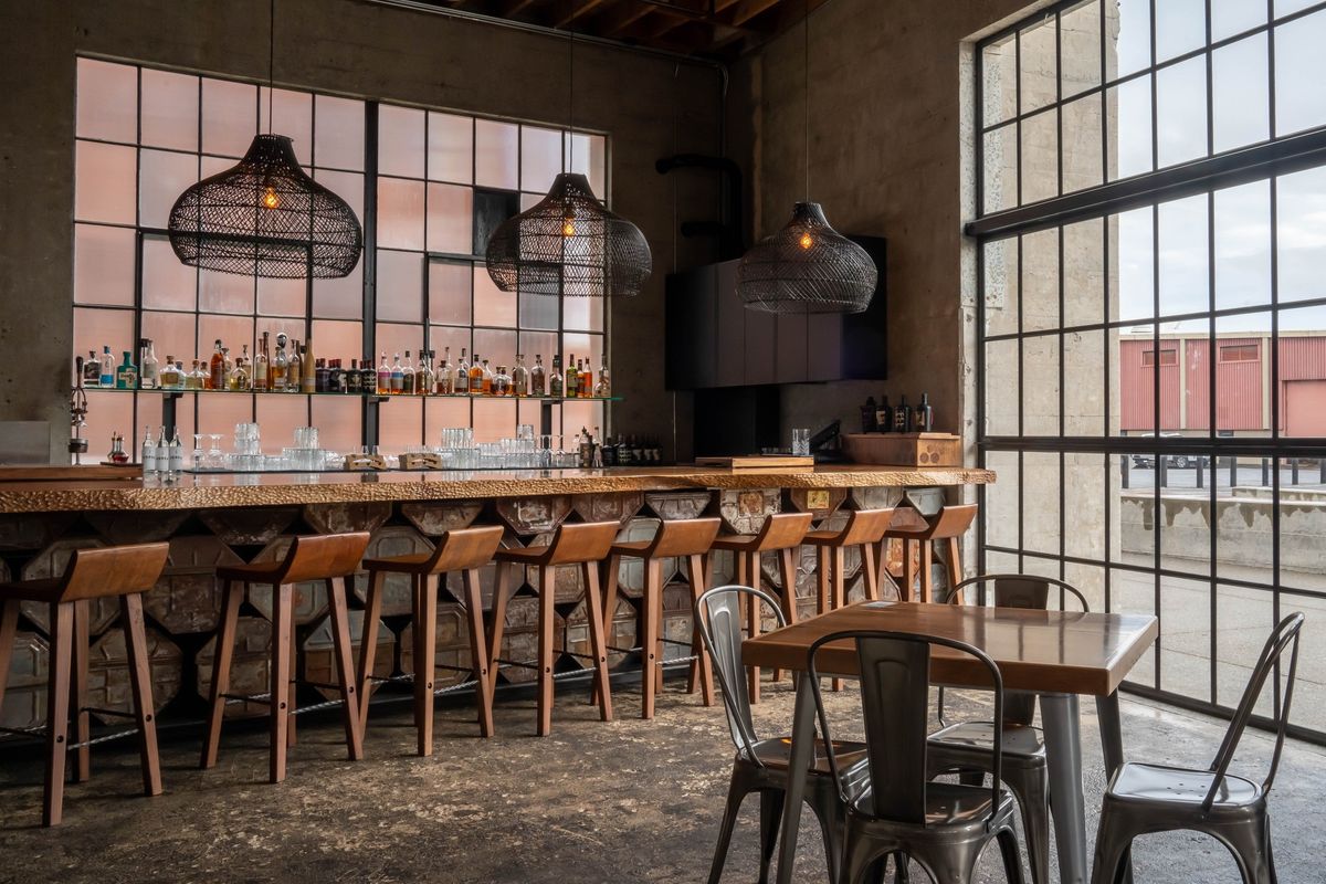 Industrial chic bar with wooden stools and metal chairs, large windows, and hanging lamps.
