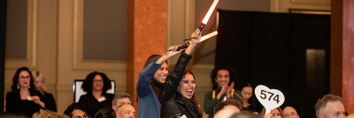 Joyful auction participants holding bid paddles and lightsabers in a crowded event hall.