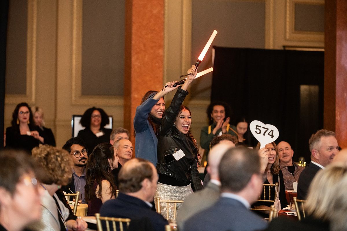 Joyful auction participants holding bid paddles and lightsabers in a crowded event hall.