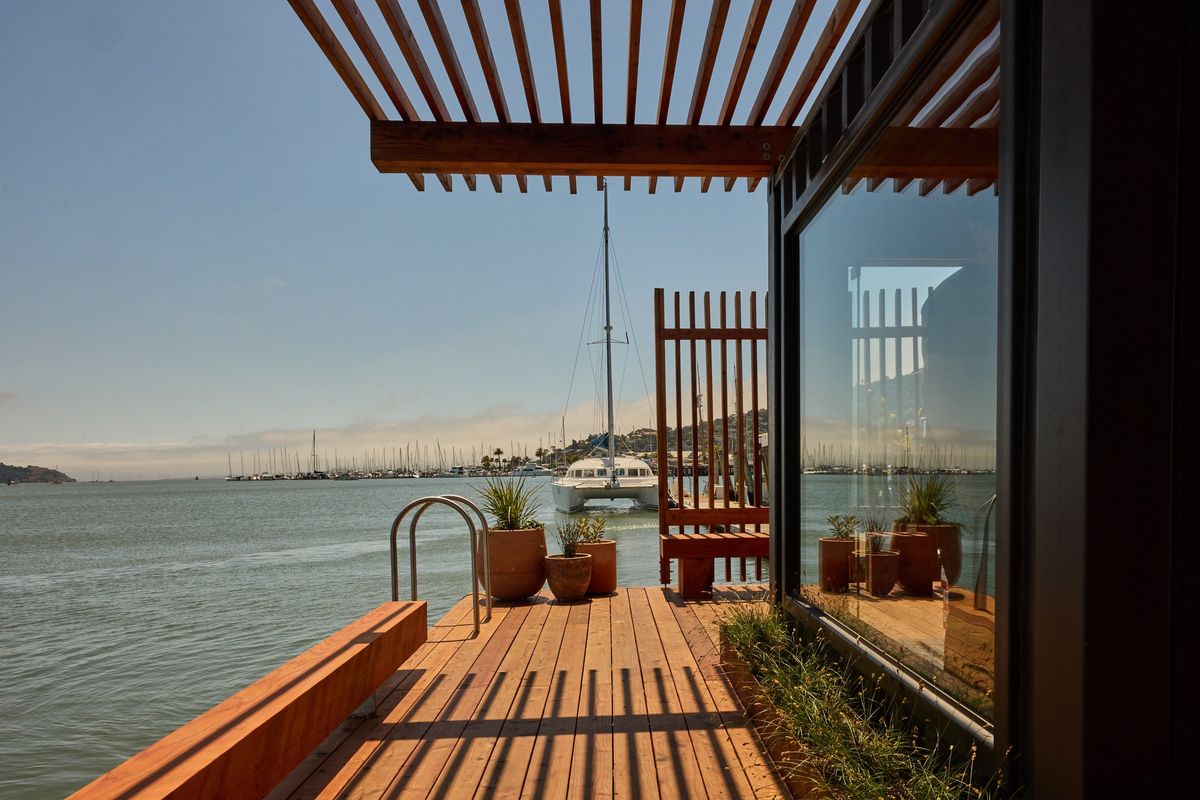 Lakeside deck with potted plants, lattice roof, and moored boats under a clear sky.