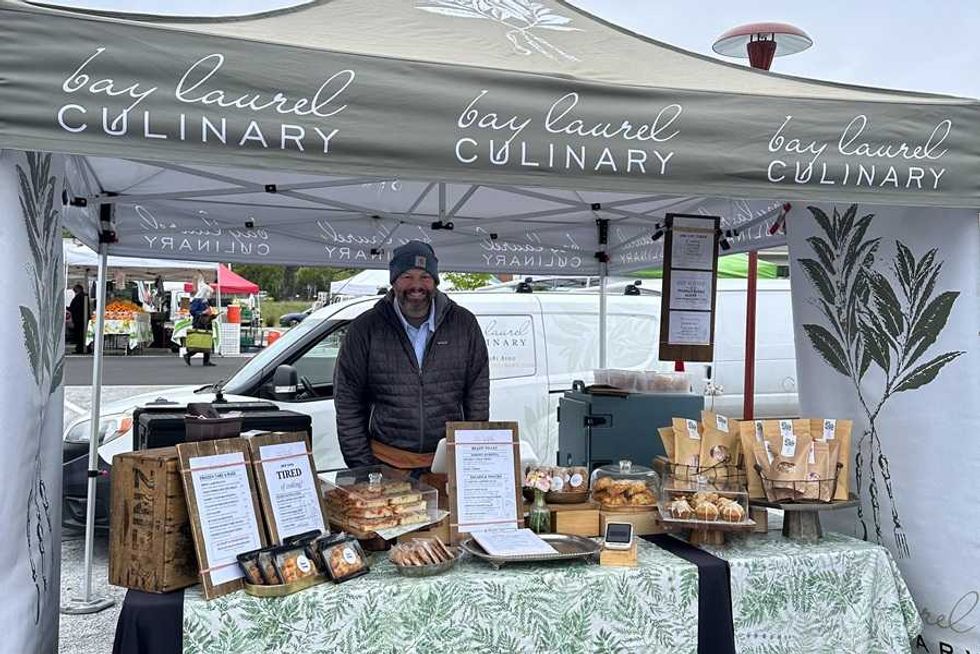 Man at Bay Laurel Culinary booth with baked goods at an outdoor market.