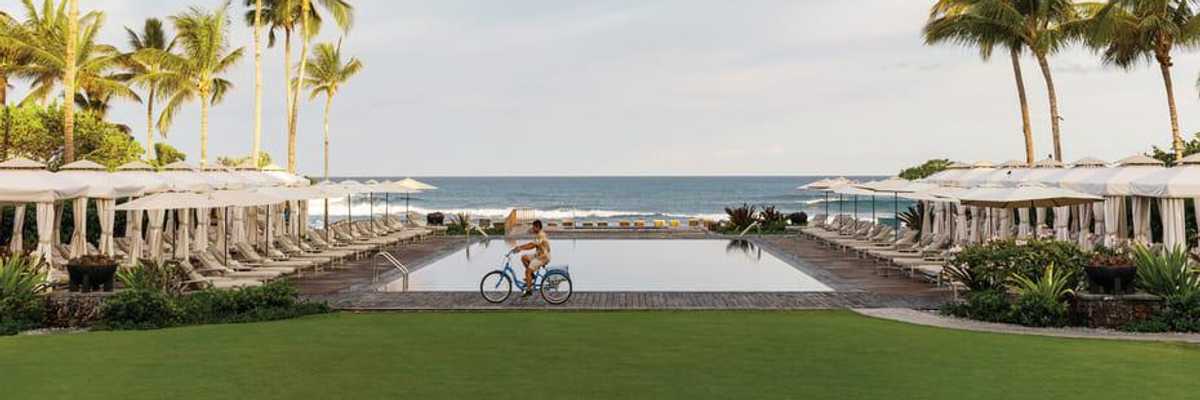 Man cycling by poolside with palm trees and ocean view.