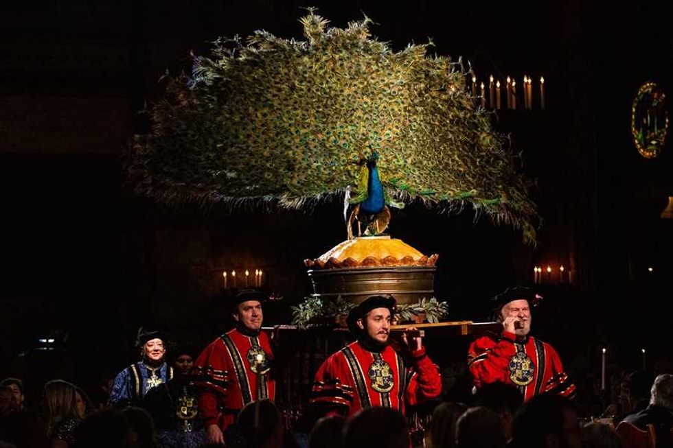 Men in red robes carrying a large peacock display in a dimly lit setting.