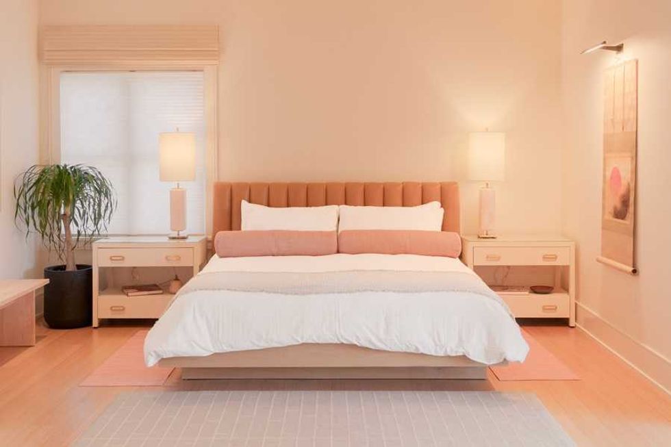 Minimalist bedroom with a cushioned bed, lamps, plant, and soft lighting.