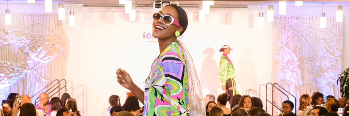 Model in colorful outfit smiles on runway; audience seated around tables.