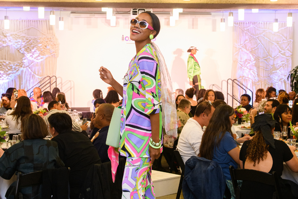 Model in colorful outfit smiles on runway; audience seated around tables.