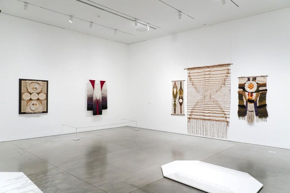 Modern art gallery with various textile wall hangings displayed on white walls.