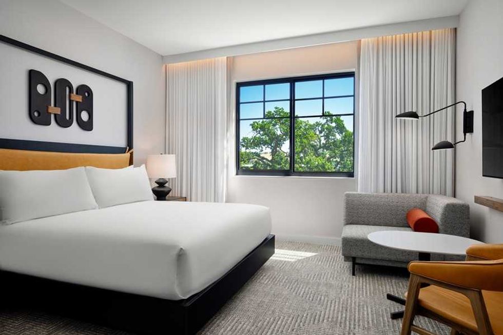Modern hotel room with large bed, gray sofa, and window view of trees.